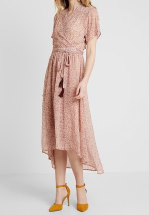 Kvinde iført en pink blomsterwrap-kjole med bundet talje og asymmetrisk kant, parret med karrygule sandaler med ankelrem.