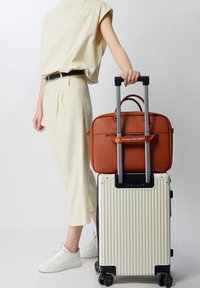 Conjunto de viaje de color crema, bolso de cuero marrón para portátil sobre una maleta rígida blanca con textura y ruedas y asas negras.