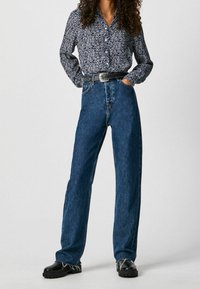 Kvinde iført en blå blomster T-shirt med knapper, stylet med højtaljede blå jeans, et sort bælte og sorte chunky sko.