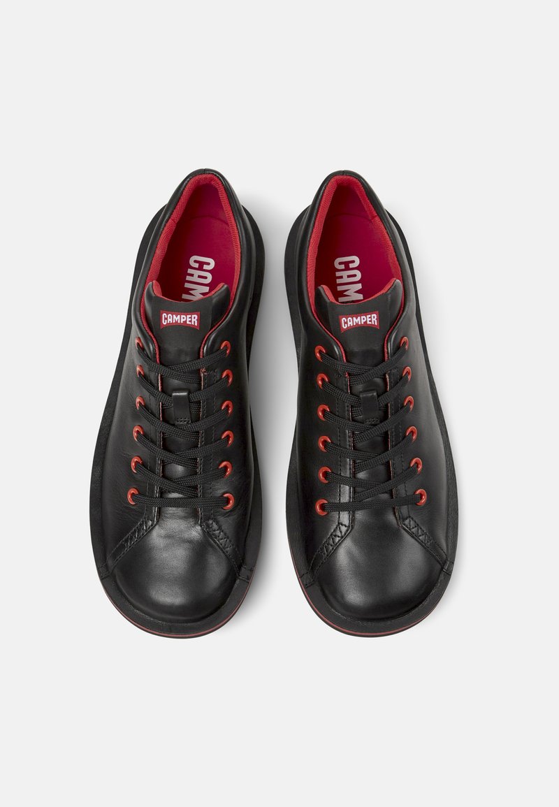 Schwarze Ledersneaker mit roten Akzenten, runde Zehenpartie, flache Schnüren und "CAMPER"-Logo auf der Zunge. Glatte Oberfläche und kontrastierendes Innenfutter.