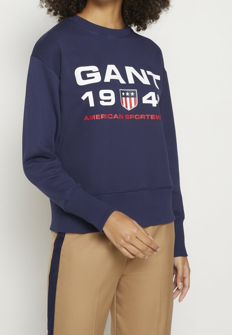 Námořnická modrá mikina z bavlny s kulatým výstřihem. Obsahuje výrazný bílý text "GANT 1944" a "AMERICAN SPORTSWEAR" v červené barvě.