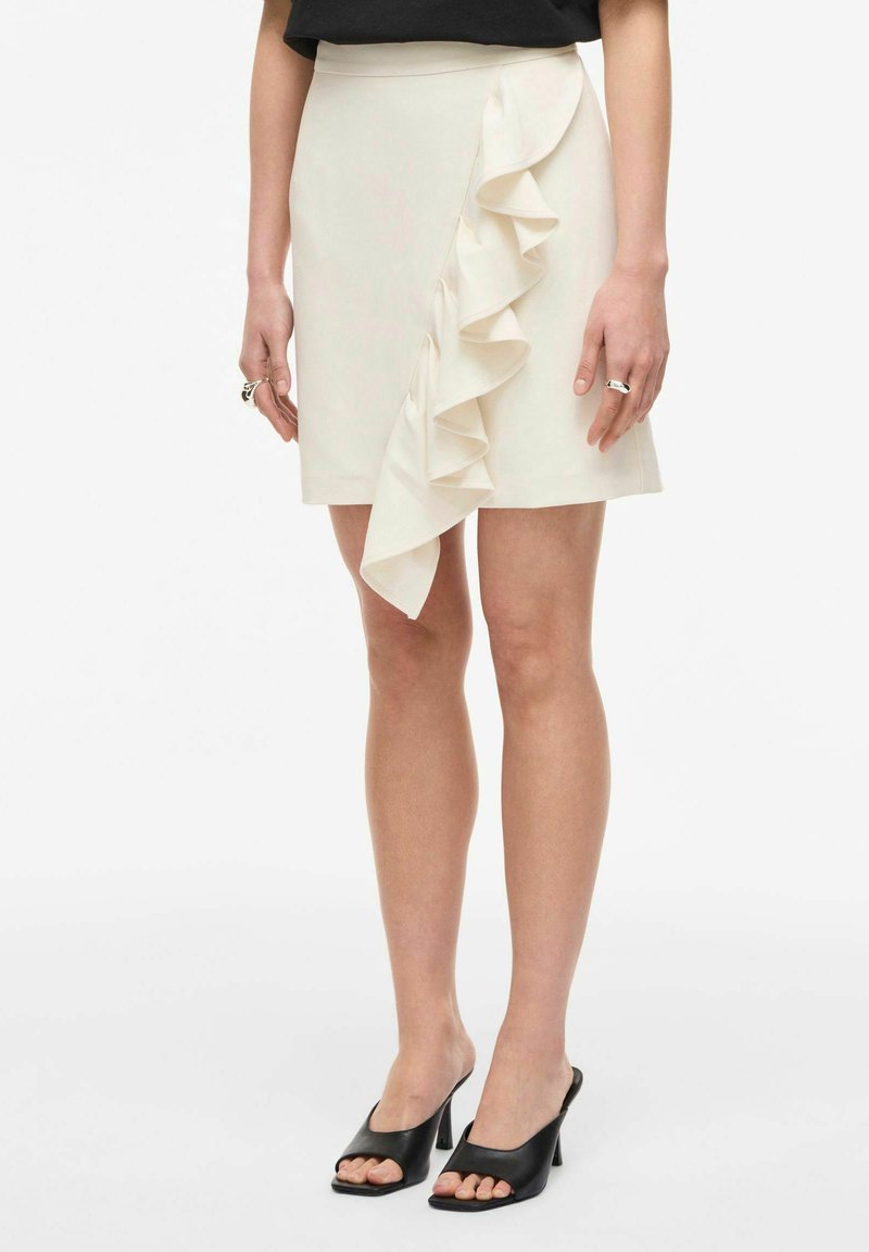 Femme portant une mini-jupe crème avec un détail de volant à l'avant et des mules à talons noires à bout ouvert, debout sur fond blanc.