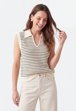 Vrouw met lang bruin haar die lacht, draagt parel oorbellen, een mouwloze crème- en zwart gestreepte gebreide top en lichtbeige broek.