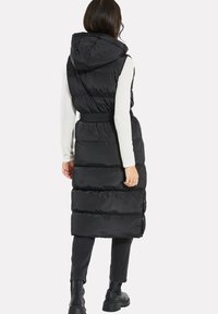 Schwarz gesteppte Weste mit Kapuze, tailliertem Schnitt und langer Silhouette; zeichnet sich durch eine gesteppte Textur und seitliche Reißverschlüsse zum Verschließen aus.