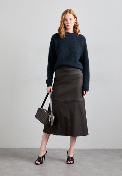 Pull en laine bleu marine, jupe en cuir noir avec un ourlet évasé, talons noirs ouverts, et sac en tissu tissé sombre avec des accents dorés.