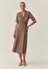Knielanges, braunes Wickelkleid mit kurzen Ärmeln, V-Ausschnitt und seitlichem Drapierungsdetail. Glatter Stoff mit lockererPassform, kombiniert mit beigen Absatzschuhen.
