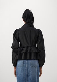 Svart peplum-jakke med puffede ermer, laget av lett stoff. Har en strukturert krage og dekorative volanger i midjen.