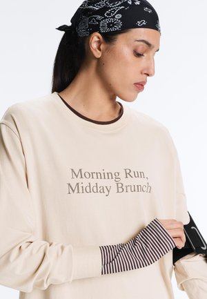 Nő krémszínű pulóvert visel "Reggeli futás, délutáni brunch" felirattal, fekete mintás kendőt és fekete karpántot a bal karján.