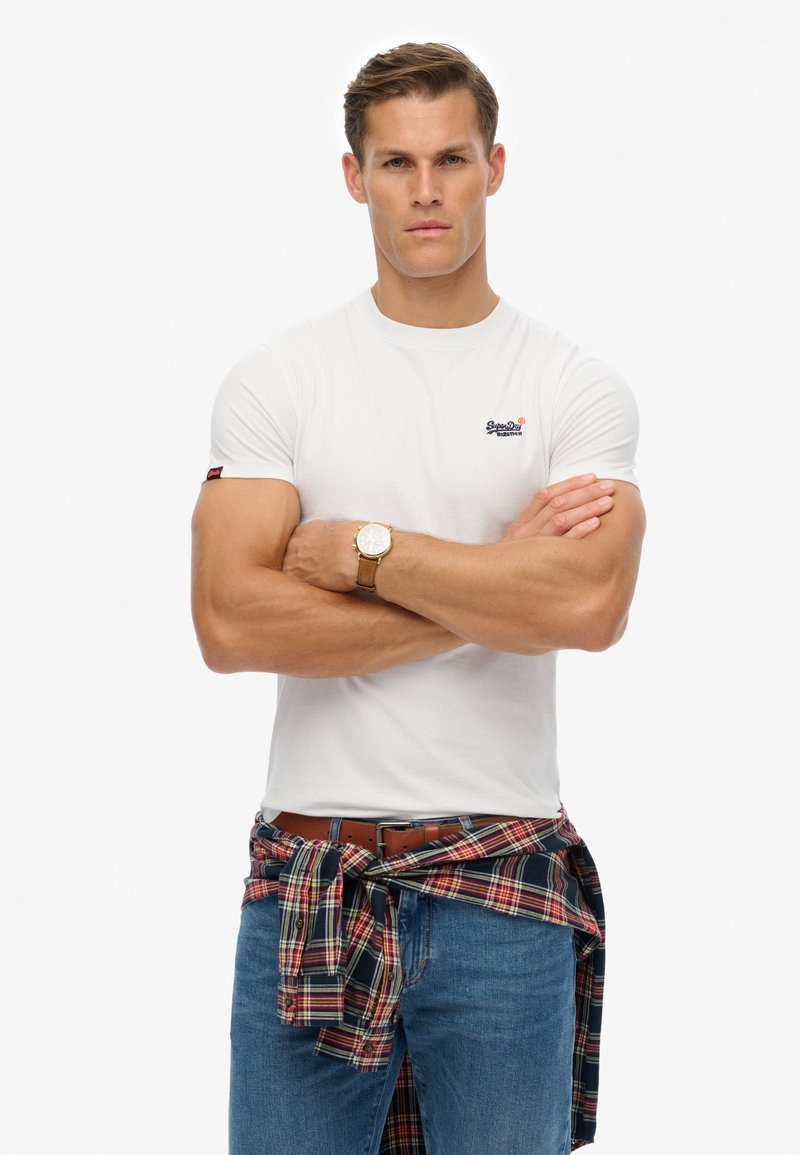 T-shirt bianco a maniche corte con logo sul lato sinistro del petto, abbinato a jeans blu e una camicia flanella a quadri legata in vita. Cintura e orologio marroni.