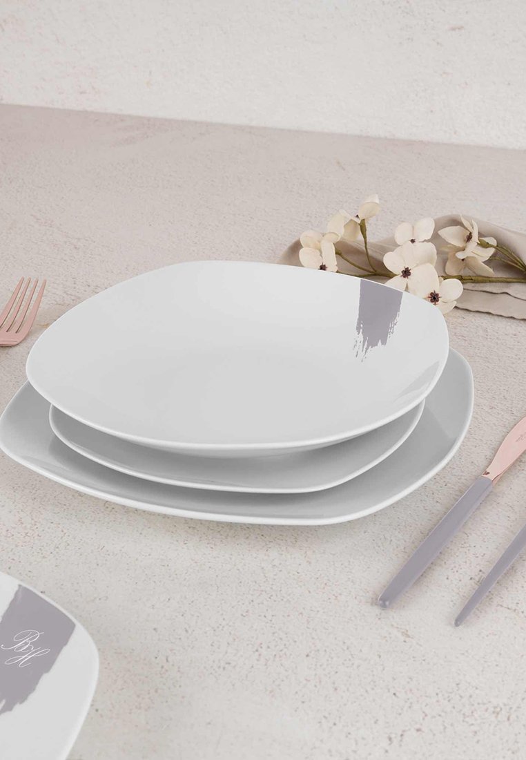 Weiße quadratische Teller übereinandergestapelt, mit einem grauen Pinselstrich-Muster. Besteck mit rosa Griffen daneben platziert. Blumendekoration in der Nähe.