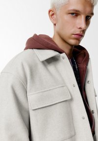 Lichtgrijze wollen jas met een kraag, voorzien van een borstzak en gelaagd met een bordeauxrode hoodie eronder. Soepele textuur.