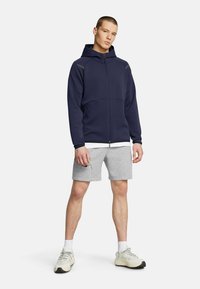Navy zip-up hoodie with hood, featuring gray accents. Paired with light gray shorts and neutral sneakers. Smooth fabric and athletic fit.