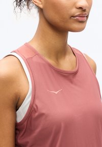 Νεαρή γυναίκα που φοράει ροζ αθλητικό τοπ χωρίς μανίκια πάνω από λευκό αθλητικό σουτιέν, το οποίο κόβεται στο ύψος του πηγουνιού και των ώμων.