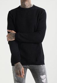 Svart stickad tröja med rund hals, texturerat mönster och ribbade ärmslut. Kombinerad med slitna gråjeans.