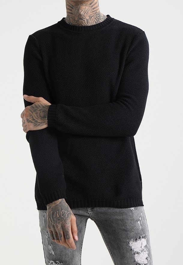 Svart stickad tröja med rund hals, texturerat mönster och ribbade ärmslut. Kombinerad med slitna gråjeans.