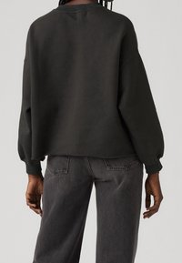 Personne portant un sweat-shirt noir ample à manches longues et un jean noir délavé, vue de dos sur un fond uni.