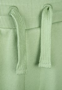Hellgrüner Stoff mit strukturiertem Finish, ausgestattet mit einem gestrickten Bund und Kordelzügen. Die Schnüre sind flach, gewebt mit einem dezenten Muster.