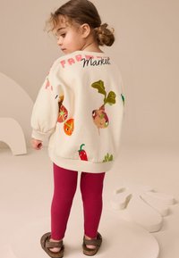 Biała bluza z kolorowymi ilustracjami warzyw, różowymi akcentami i napisem "Market". Połączona z czerwonymi legginsami i brązowymi sandałami.