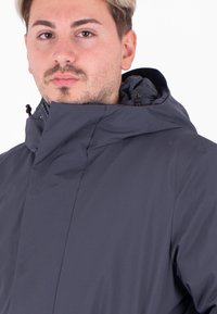 Giacca impermeabile grigio scuro con colletto alto, cappuccio regolabile e texture liscia. Presenta cordini e un design minimalista.