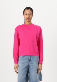 Sudadera fucsia con cuello redondo y puños acanalados, con un logo sutil en el pecho y un ajuste relajado. Combinada con pantalones cortos de mezclilla claros.