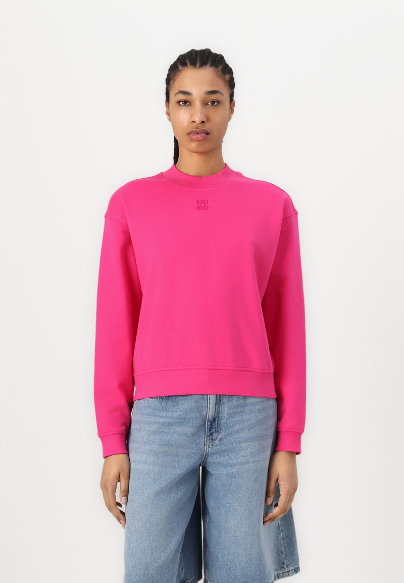 Sudadera fucsia con cuello redondo y puños acanalados, con un logo sutil en el pecho y un ajuste relajado. Combinada con pantalones cortos de mezclilla claros.