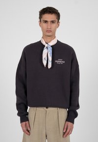 Sweat-shirt gris foncé avec poignets côtelés, présentant une broderie "PROHIBITED Racing Club" sur la poitrine. Porté avec une écharpe claire à motifs.