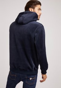 Sudadera azul marino con un acabado suave y texturizado. Cuenta con una capucha grande, puños y dobladillo de canalé, complementada con jeans de mezclilla oscura.