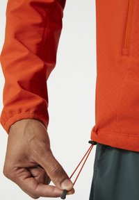 Chaqueta roja con un acabado texturizado, que cuenta con un bolsillo con cremallera y cordones ajustables en el dobladillo. Mano agarrando el cordón.