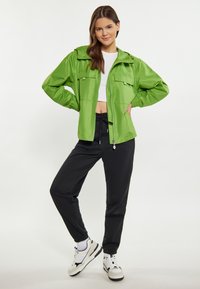 Grøn letvægtsjakke med lynlås, to frontlommer og elastiske mansjetter, kombineret med sorte sweatsbukser og hvide sneakers.