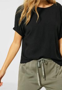 Top corto nero a maniche corte con maniche arricciate, abbinato a pantaloni verde oliva chiaro con cintura regolabile e dettagli stampati sui legacci.