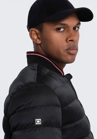 Giacca a vento nera con design trapuntato, colletto a righe rosse e bianche e patch con logo sulla manica sinistra. Accompagnata da un berretto nero.