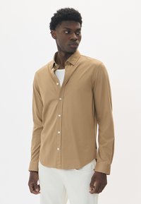 Svetlohnedá košeľa s dlhým rukávom vyrobená z hladkej tkaniny, s zapínaním na gombíky, špicatým golierom a bielymi gombíkmi.