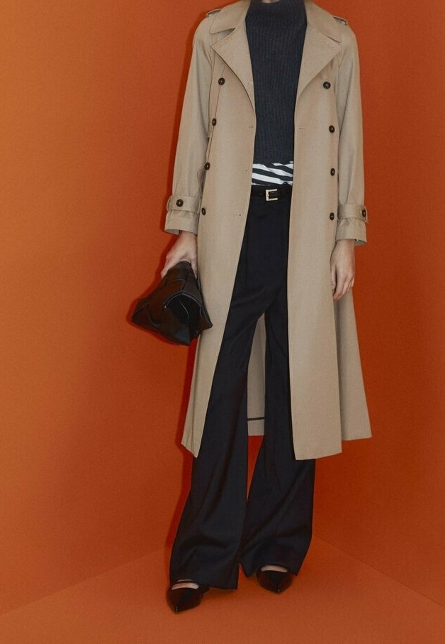 Personne portant un trench beige, un col roulé noir, un pantalon large noir, tenant un sac noir sur un fond orange.