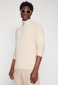 Pulover din fleece de culoare crem, cu guler înalt și fermoar la jumătate. Prezintă un material texturat și mâneci ajustate. Purtați-l cu pantaloni bej.