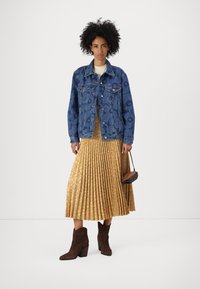 Denim jakna s cvetnim vzorcem, gorčična plisirana krila s krožnimi tiskanji, rjavi antilopski gležnjarji, majhna torbica v roki.