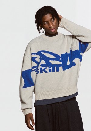 Personne aux cheveux foncés tressés portant un pull beige avec un grand motif de texte abstrait bleu et un pantalon noir, posant avec la main derrière la tête.