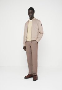 Άνδρας με σκουρόχρωμο δέρμα που φοράει μπεζ σακάκι, κρεμ φούτερ, ανοιχτό καφέ παντελόνι και καφέ δερμάτινα παπούτσια, στέκεται μπροστά σε λευκό φόντο.