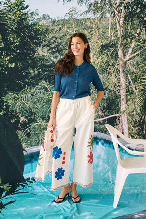 Uśmiechnięta kobieta, ubrana w niebieską koszulkę z krótkim rękawem oraz białe spodnie o szerokich nogawkach z kolorowymi kwiatowymi nadrukami, stoi przy basenie, w tle drzewa.