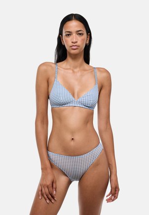 Jeune femme aux longs cheveux foncés portant un bikini à carreaux bleu et blanc sur un fond clair uni.