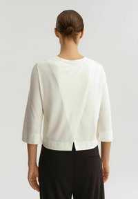 Top bianco, corto, con maniche a tre quarti, caratterizzato da un design posteriore a forma di V e tessuto liscio. Indossato sopra pantaloni neri.