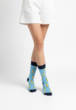 Kājas baltos šortos un spilgti zilās zeķēs ar dzelteniem meerkatu rakstiem, stāvot uz balta fona.