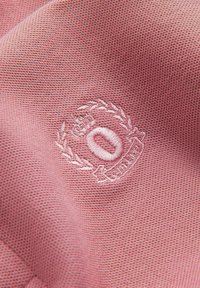 Broderad logotyp på strukturerad rosa tyg, med ett runt emblem med lageraccent och en krona, som visar bokstaven "O."