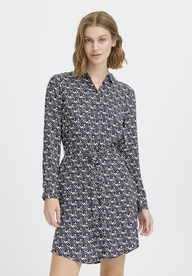 Moteris vilkinti tamsiai mėlyną raštuotą ilgos rankovės suknelę su dirželiu, stovinti prieš paprastą šviesų foną.