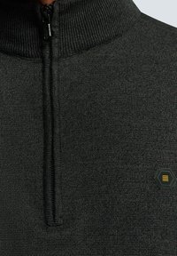 Zwarte zip-up trui met ribbelkraag, gemaakt van een textuur gebreide stof en voorzien van een klein hexagonale logo in de onderste hoek.