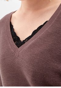 Pull violet en maille avec un col en V, dévoilant un caraco en dentelle noire en dessous. La texture est côtelée avec une finition douce.