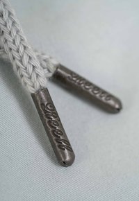 Cordones trenzados de color gris claro con terminales metálicos. Los terminales están grabados con "Suerlo" y "Dieel", destacando los detalles de la marca.
