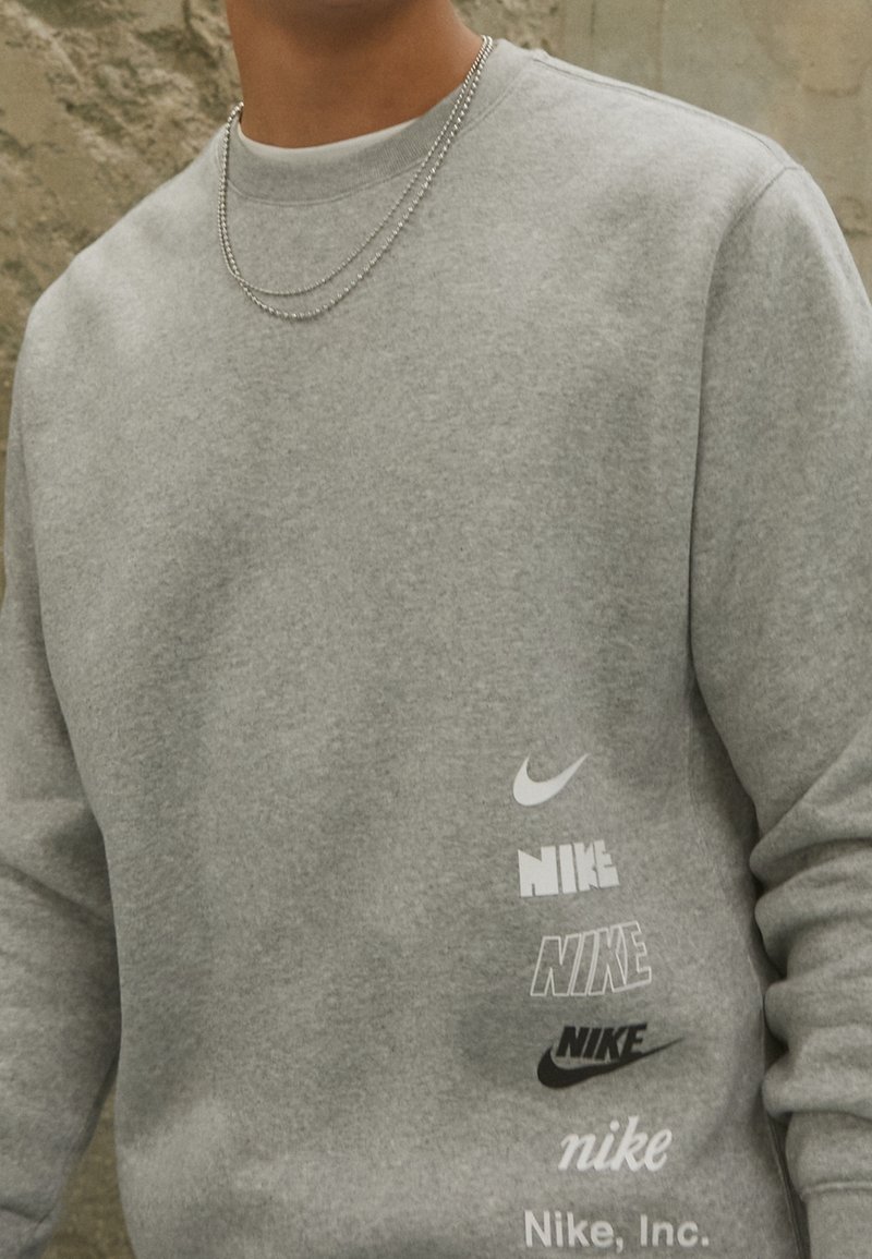 Šedá mikina s kulatým výstřihem, na přední straně s různými logy Nike v kontrastních barvách a hladkým, jemným povrchem.