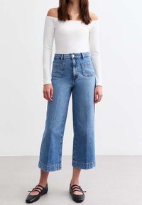 Jeans de pierna ancha de talle alto en denim azul claro, con bolsillos delanteros y dobladillo desflecado. Combinados con un top blanco de hombros descubiertos y zapatos planos negros.