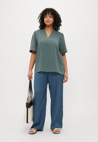 Αδειανό μπλουζάκι σε teal με κοντά μανίκια και V-λαιμόκοψη σε υφή, σε συνδυασμό με φαρδιά μπλέ παντελόνια και μια καμβά τσάντα tote.