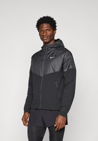 Czarna sportowa kurtka z błyszczącą czarną górną częścią, kapturem, zapięciem na zamek, bocznymi kieszeniami oraz logo Nike na piersi.